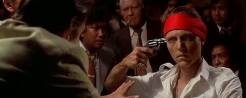 Scen från filmen Dead Hunter - en man i röd bandana håller en pistol mot sitt huvud med massor av människor runtom